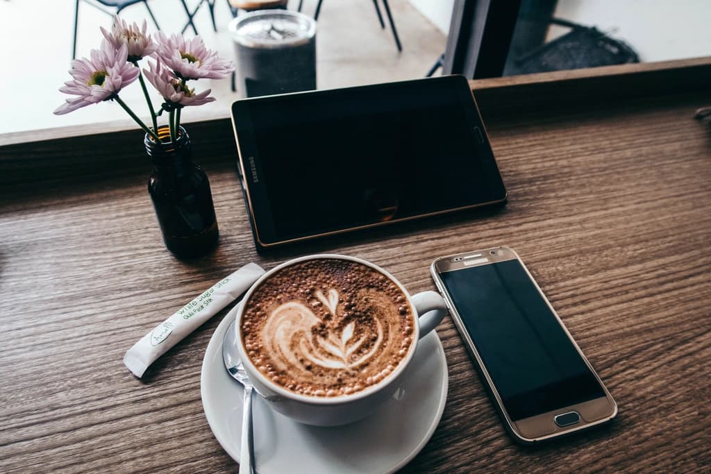 Un téléphone et une tablette posés sur une table de café, à côté d’un cappuccino.