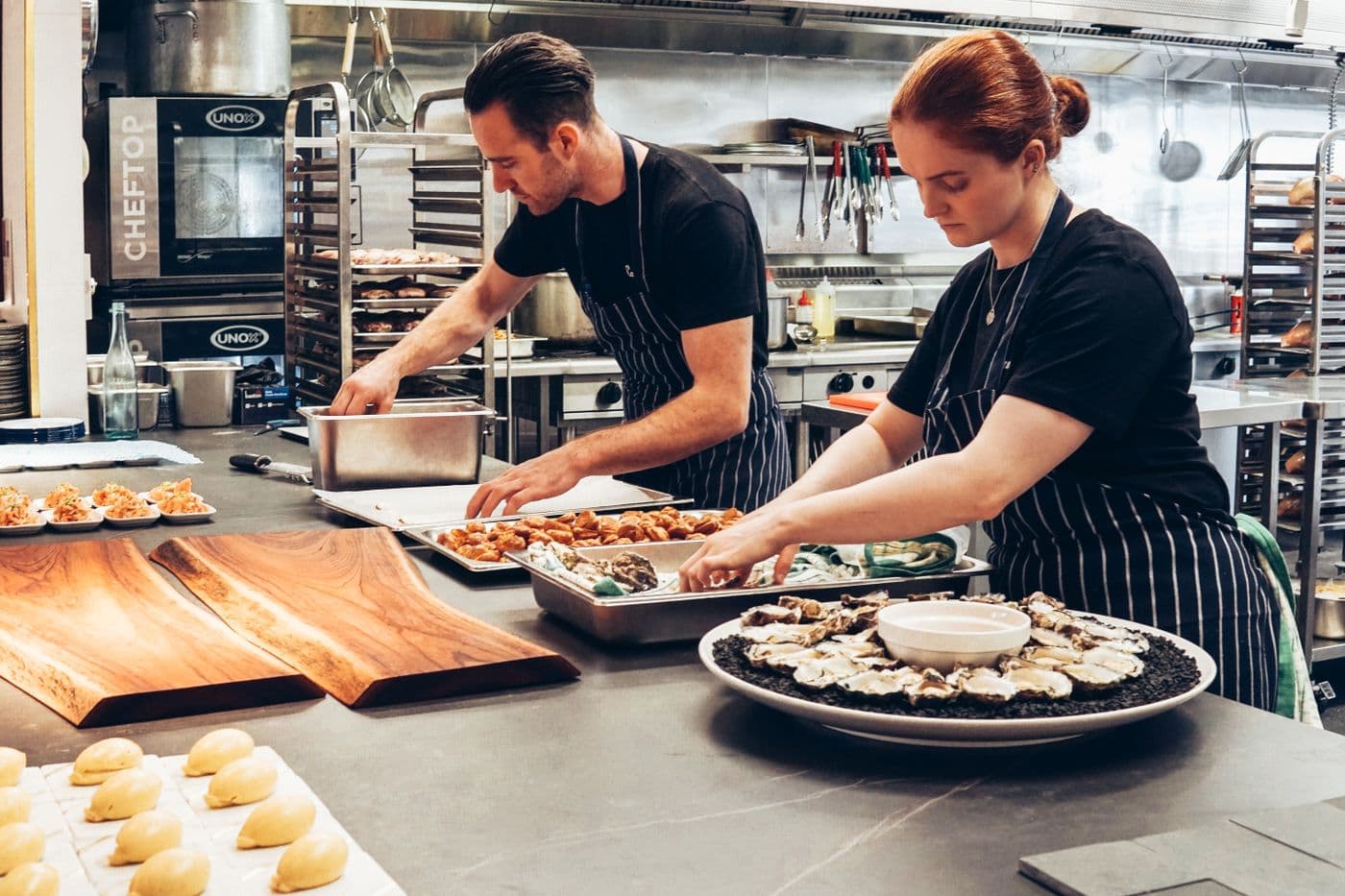 Deux cuisiniers dressent des assiettes à des postes distincts, dans une cuisine professionnelle en plein service.